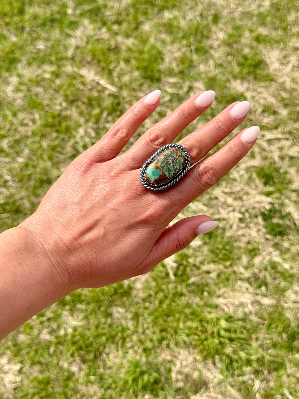 Hand wearing a large ring with a turquoise stone against a grassy background