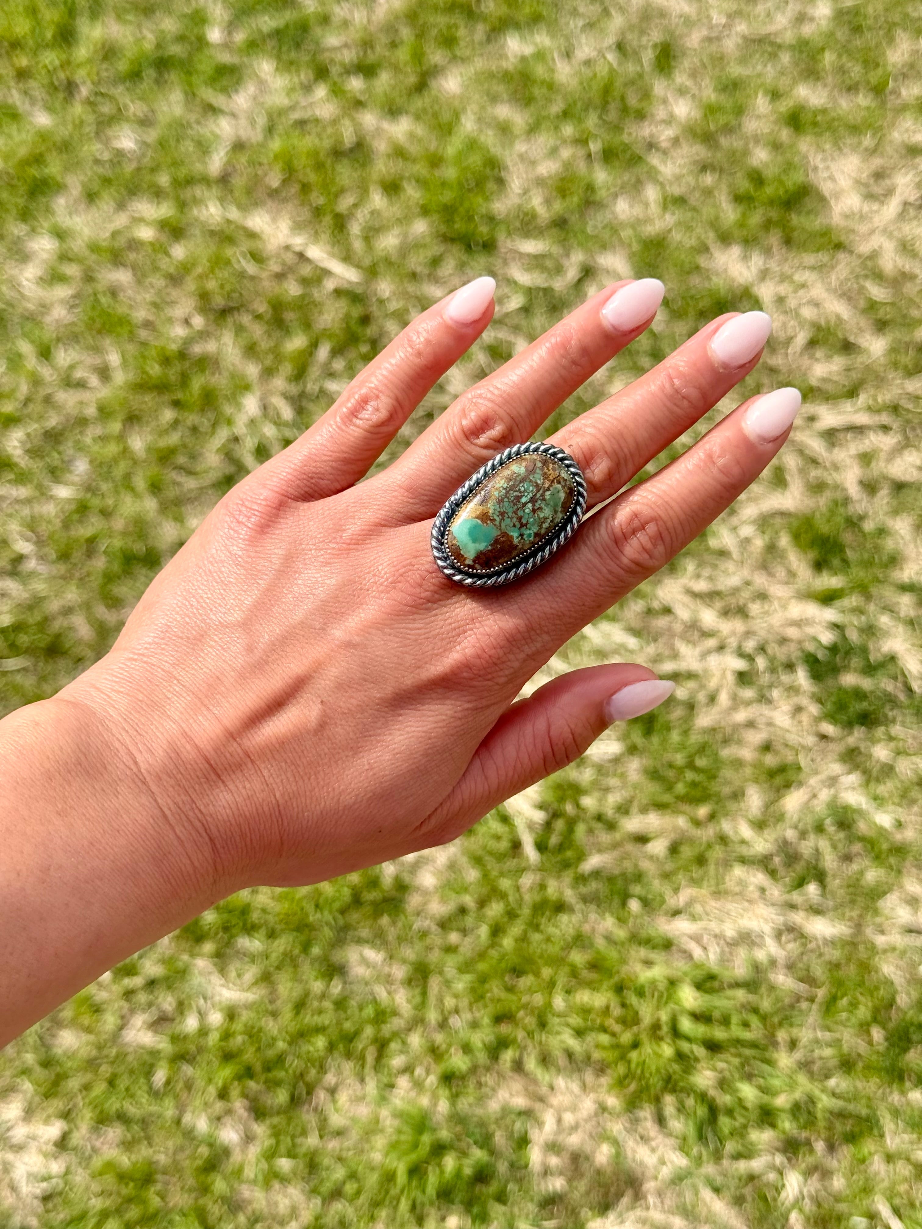 Hand wearing a large ring with a turquoise stone against a grassy background