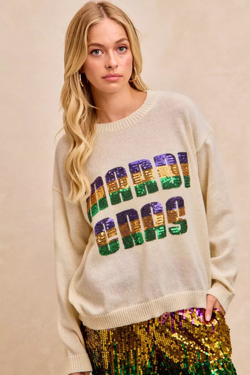 Ivory BiBi sweater knit top with sequin Mardi Gras lettering in purple, gold, and green, soft stretch knit fabric, crew neckline, short sleeves, relaxed fit, festive seasonal sweater top